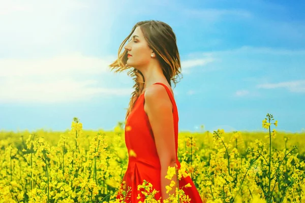 Joven hermosa chica en un vestido rojo de cerca en el medio del campo amarillo con flores de rábano y sunligh —  Fotos de Stock