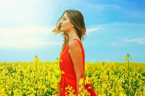 Young beautiful girl in a red dress close up in the middle of yellow field with radish flowers and sunligh — Stock Photo, Image