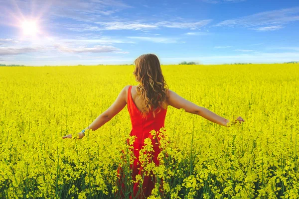 Menina bonita nova em um vestido vermelho perto no meio do campo amarelo com flores de rabanete e sol — Fotografia de Stock