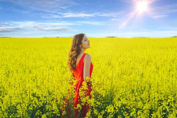 Menina bonita nova em um vestido vermelho perto no meio do campo amarelo com flores de rabanete e sol — Fotografia de Stock