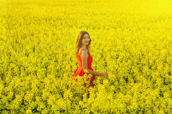 Young beautiful girl in a red dress close up in the middle of yellow field with radish flowers and sunligh — Stock Photo, Image