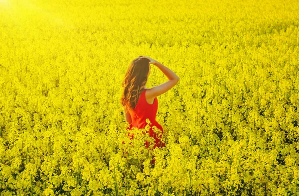 Menina bonita nova em um vestido vermelho perto no meio do campo amarelo com flores de rabanete e sol — Fotografia de Stock