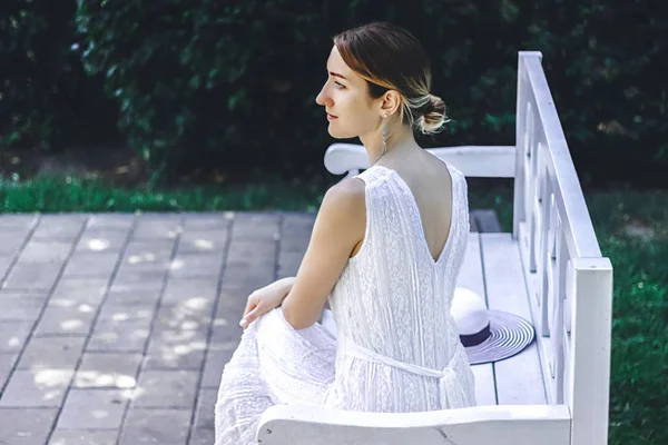 Young beautiful girl in a white dress sitting on a white bench in the summer park — Stock Photo, Image