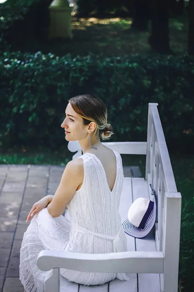 Young beautiful girl in a white dress sitting on a white bench in the summer park — Stock Photo, Image