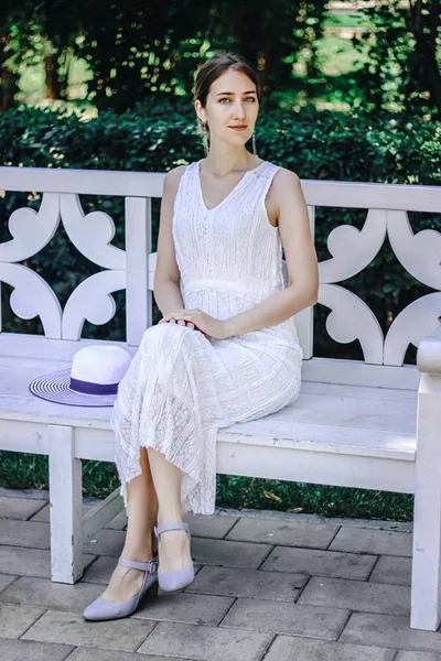 Mladá krásná dívka v bílých šatech, sedící na bílé lavičce v letním parku — Stock fotografie