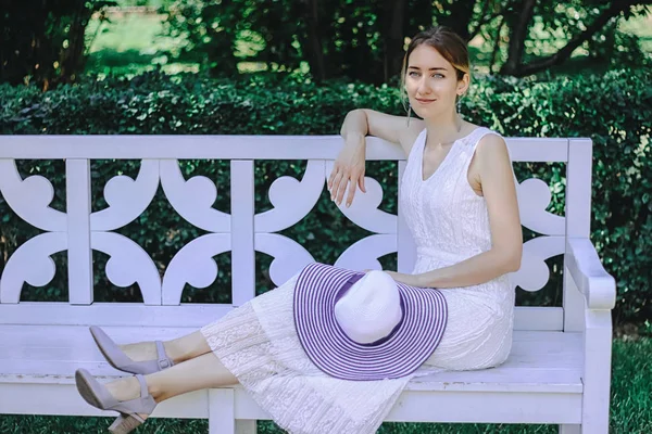 Mladá krásná dívka v bílých šatech, sedící na bílé lavičce v letním parku — Stock fotografie