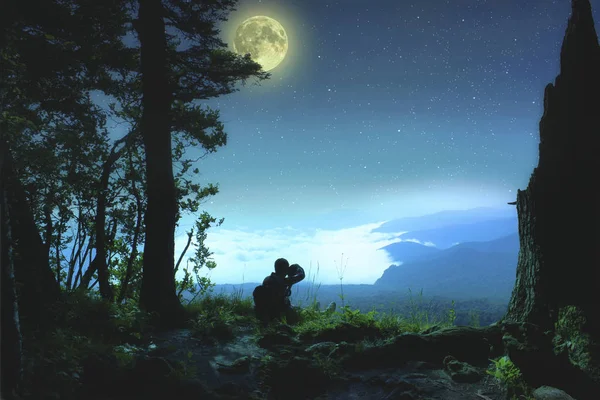 Silueta de un joven que se sienta en el borde de la plataforma de observación y disfruta del paisaje nocturno con cielo estrellado y luna llena — Foto de Stock