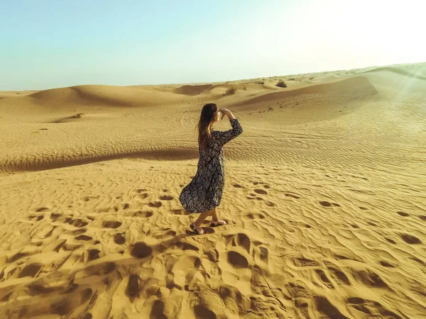 Young beautiful woman in long dress and with a hat in the middle of the Dubai desert with sunlight — Stock Photo, Image