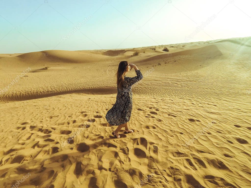 Young beautiful woman in long dress and with a hat in the middle of the Dubai desert with sunlight