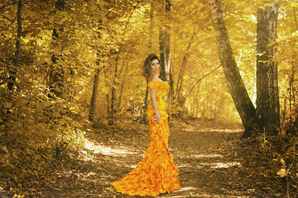 Young beautiful girl in a dress made of autumn leaves in the park in autumn season. Art photo.