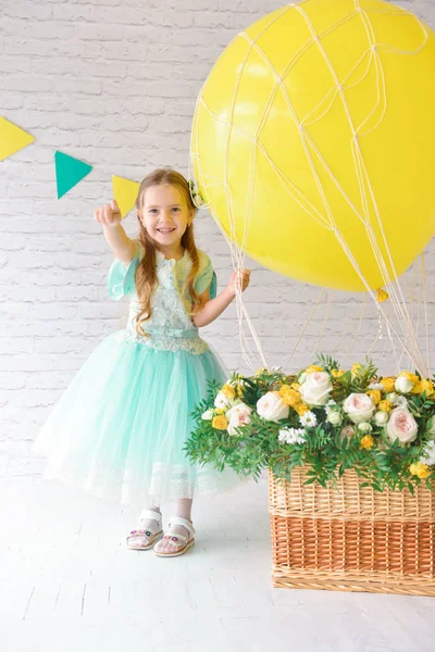 A girl in an elegant dress is standing in the studio next to a large balloon and a basket of flowers. Festive decor, birthday decorations