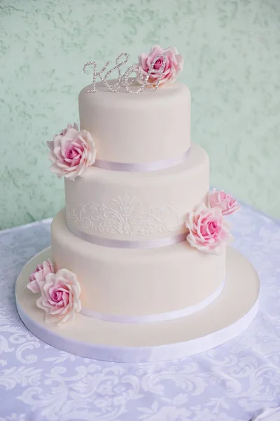 Τριών επιπέδων γαμήλια τούρτα με ροζ τριαντάφυλλα από μαστίχα στέκεται πάνω στο τραπέζι μια λεπτή υπόβαθρο. γράμματα νύφη και το γαμπρό — Φωτογραφία Αρχείου