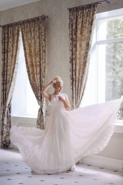 Das blonde Mädchen auf dem Bild der Braut steht in einem fliegenden hellen Kleid am Fenster. Hochzeitsfoto. Sanft ruhiges Bild. Geringes Rauschen — Stockfoto