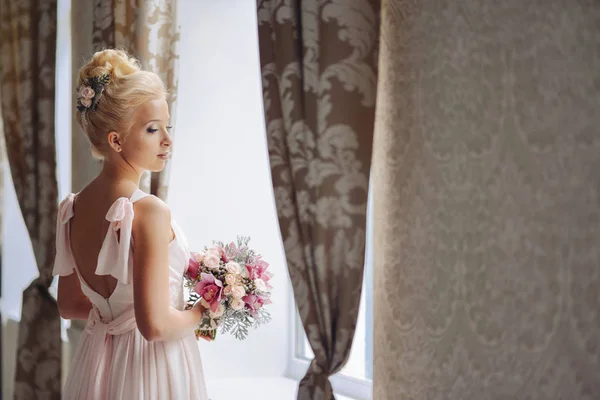 Красивая молодая женщина стоит в образе невесты со свадебным букетом роз и орхидей, стоящих у окна в платье с обнаженной спиной — стоковое фото