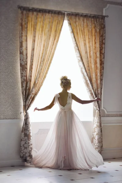 Das blonde Mädchen auf dem Bild der Braut steht in einem fliegenden hellen Kleid am Fenster. Hochzeitsfoto. Sanft ruhiges Bild. Geringes Rauschen — Stockfoto