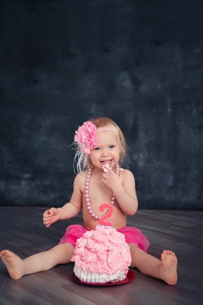 Thema verjaardag voor een pret emotionele meisje van de blonde smash de taart in roze kleur op een zwarte achtergrond. gestileerde foto sessie traditie met zoete decor en ballonnen — Stockfoto