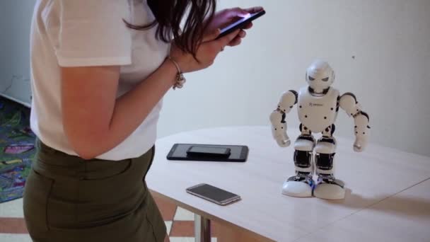 Wit-Rusland, Minsk, robot tentoonstelling, 3 juni 2019: vrouw besturingselementen van de telefoon sets commando's witte plastic robot dansen op tafel binnenshuis — Stockvideo