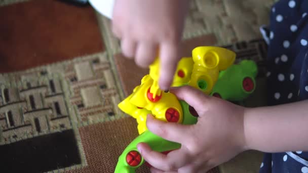 El niño juega en el interior en un sofá con el kit de construcción de diseño en forma de dinosaurio. Los dedos de los niños recogen un juguete rompecabezas de color brillante con un destornillador . — Vídeos de Stock