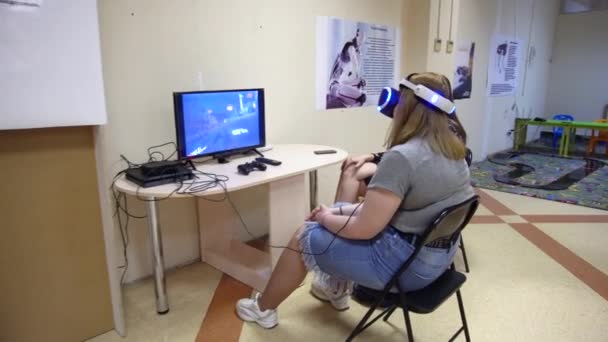 Wit-Rusland, Minsk, robot tentoonstelling, 3 juni 2019: jong meisje zittend op een stoel in de kamer voor de monitor het dragen van een bril van Virtual Reality speelt games Racing Cars — Stockvideo