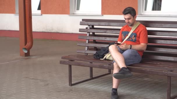 Homem entediado sentado no banco do lado de fora esperando e chamando em um telefone que não tem resposta — Vídeo de Stock