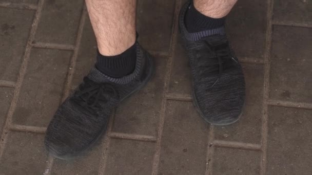 Gambe maschili di uomo seduto in scarpe da ginnastica nere stanno bussando sull'asfalto in previsione di qualcosa — Video Stock