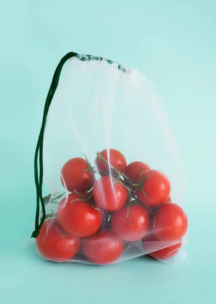 Ein reifer Zweig einer Kirschtomate liegt in einem Mehrwegbeutel auf einem isolierten blauen Hintergrund. Umgang mit Einweg-Plastiktüten Stockbild