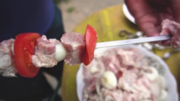 Kebabs koken. Close-up mannelijke hand snaren stukken vlees op spiesje op een zomerdag buiten. gemarineerde vlees en tomaten doorboord op een houten achtergrond een rustieke levensstijl — Stockvideo