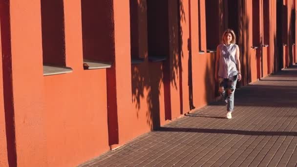 Glückliche junge Frau Blondine geht vor dem Hintergrund der roten Wand der städtischen Hintergrund. in lässiger Kleidung zerrissene Jeans und Hemd — Stockvideo
