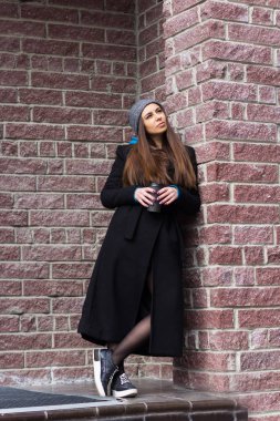 şık genç kadın renksizdir uzun siyah ceket ve duvara karşı gitmek için kahve ile gri örgü şapka