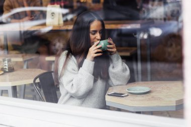 genç güzel şirin esmer kız café throuth cam pencere bir masada kahve içme gri süveterli portresi