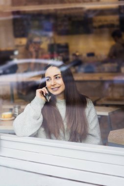 genç güzel gülümseyen esmer kız gri süveterli café throuth cam pencere bir masada telefonda konuşan portresi.