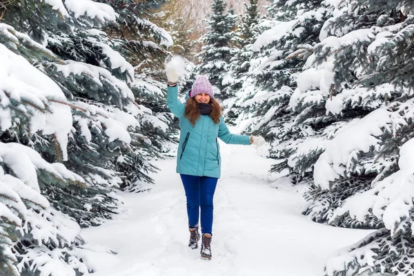 young happy excited woman in knitted hat and blue down jacket throws snow in spruce forest, winter happy portrait, holiday Christmas vibes