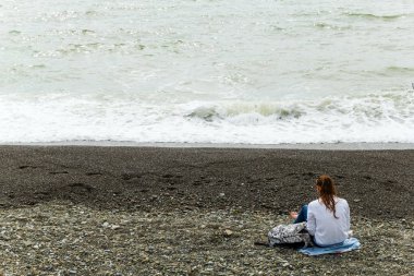 Soçi,Rusya, 20 Nisan 2019 - yalnız genç kız sahilde kameraya geri oturan ve denize bakarak
