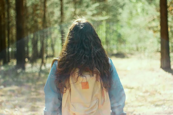 brunette woman with backpack and denim jacket stands with her back to the camera in the woods,selective focus,blurred