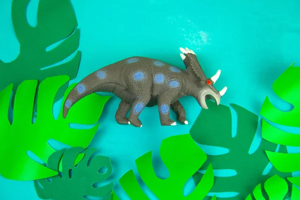 funny dinosaur toy on blue background with green paper cut tropical leaves