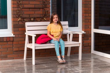 bir tuğla duvarın önünde bir bankta oturan bir sırt çantası ile güzel küçük kız