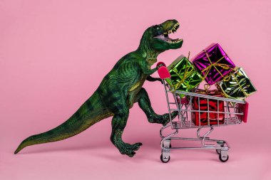 Komik yeşil dinozor oyuncağı. Alışveriş arabası dolusu hediye kutusu ve yumuşak pembe arka planı var.