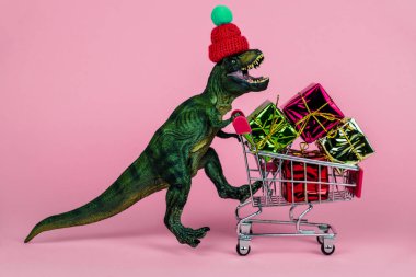 Küçük örgü şapkalı komik plastik dinozor hediye kutularıyla dolu pembe arka planda alışveriş konseptini kullanıyor.