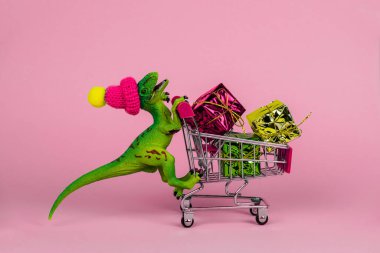 Küçük örgü şapkalı komik plastik dinozor hediye kutularıyla dolu pembe arka planda alışveriş konseptini kullanıyor.