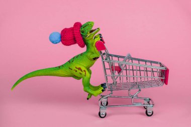 Komik plastik oyuncak dinozor minik örgü şapkalı ve pembe arka planda boş alışveriş arabası kullanıyor.