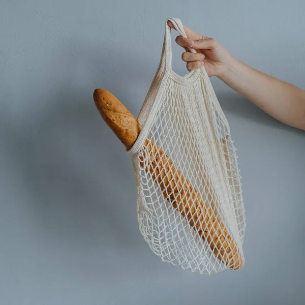female hand holding string bag with fresh baguette on a gray background