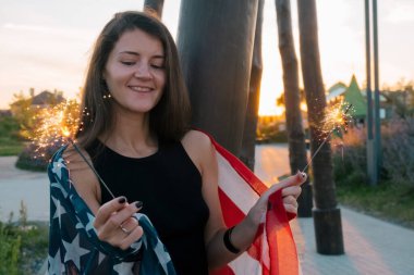 Parkta gün batımında elinde maytap ve Amerikan bayrağı resmi olan mutlu, mutlu, gülümseyen genç kadın.