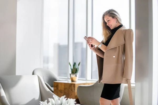 スマートフォン テキストとチャットを保持し ウィンドウ内のスカイラインと高級オフィスビルのインテリアのプロのビジネス女性 — ストック写真