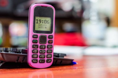 Eski düğme cep telefonu pembe rengi yakınındaki bir bilgisayar klavye masasında. Saat ve siyah beyaz ekran göstergeleri.