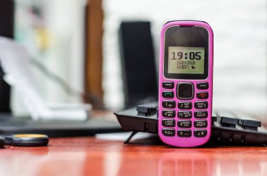 Eski düğme cep telefonu pembe rengi yakınındaki bir bilgisayar klavye masasında. Saat ve siyah beyaz ekran göstergeleri.