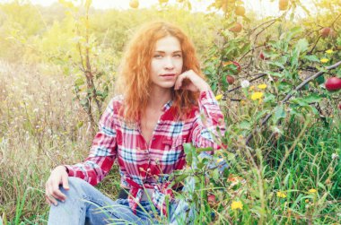 Genç kadın bir elma bahçesinde yeşil çimenlerin üzerinde oturuyor. Harv