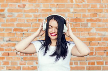 Tuğla duvar arka plan üzerinde gülümseyen bir esmer kız portresi. Beyaz kulaklık müzik dinlemek. Gençlik Kültür, albüm kapağı.