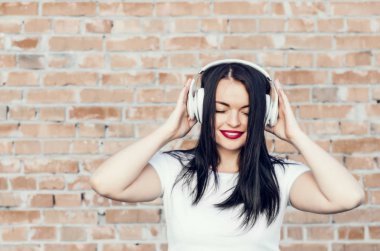 Tuğla duvar arka plan üzerinde gülümseyen bir esmer kız portresi. Beyaz kulaklık müzik dinlemek. Gençlik Kültür, albüm kapağı.