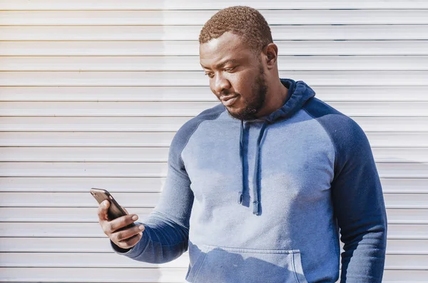 Portrait of a black guy close-up in a sports sweater on the background of a striped bright wall. Looking at phone screen. Cheerful positive person. City photo, space. Youthful culture.