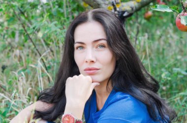 Bahçede bir elma ağacı altında yeşil çimenlerin üzerinde oturan çekici esmer kız. Doğa üzerinde bekletin. Kadın sağlığı. Yaz anıları. Cinsellik, kadınlık